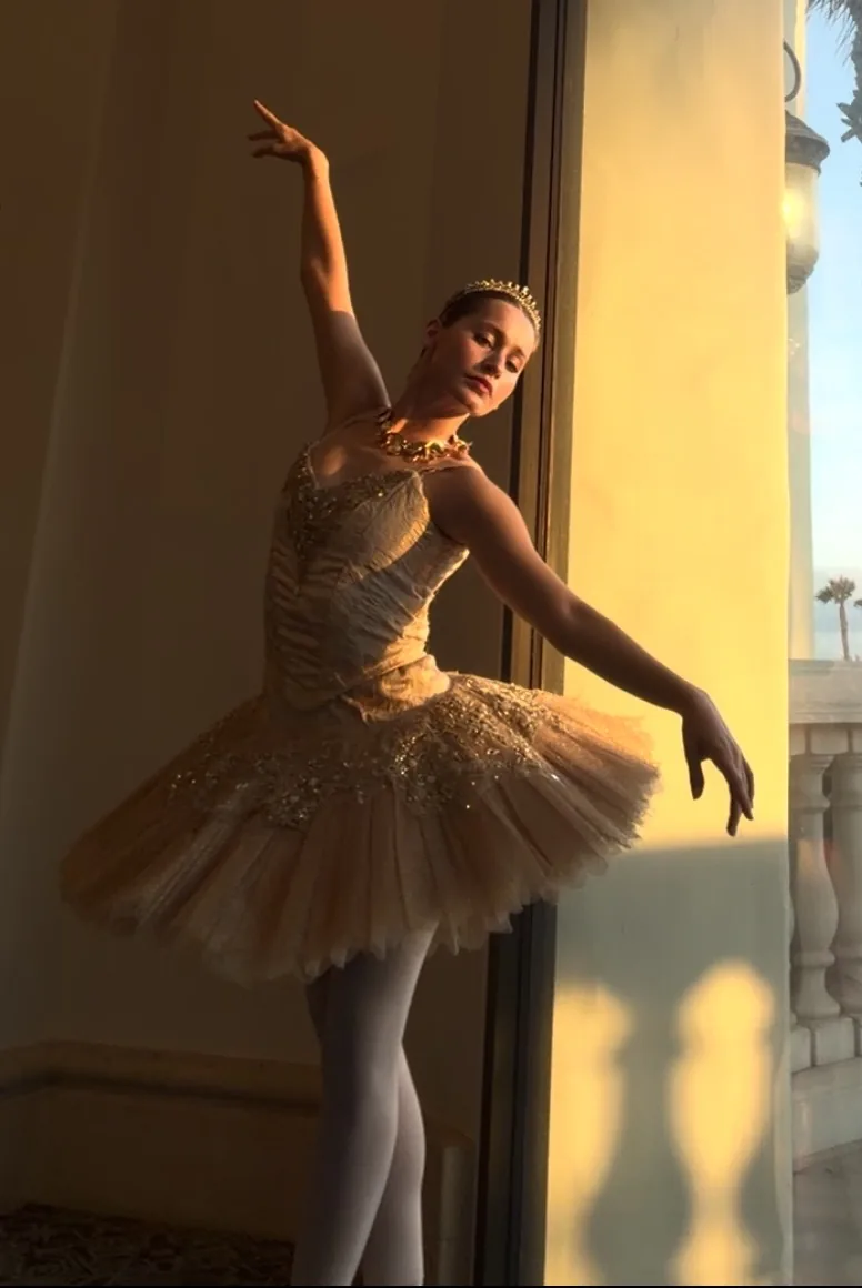 Marit in a ballet costume, posing with one arm raised beside a sunlit window.